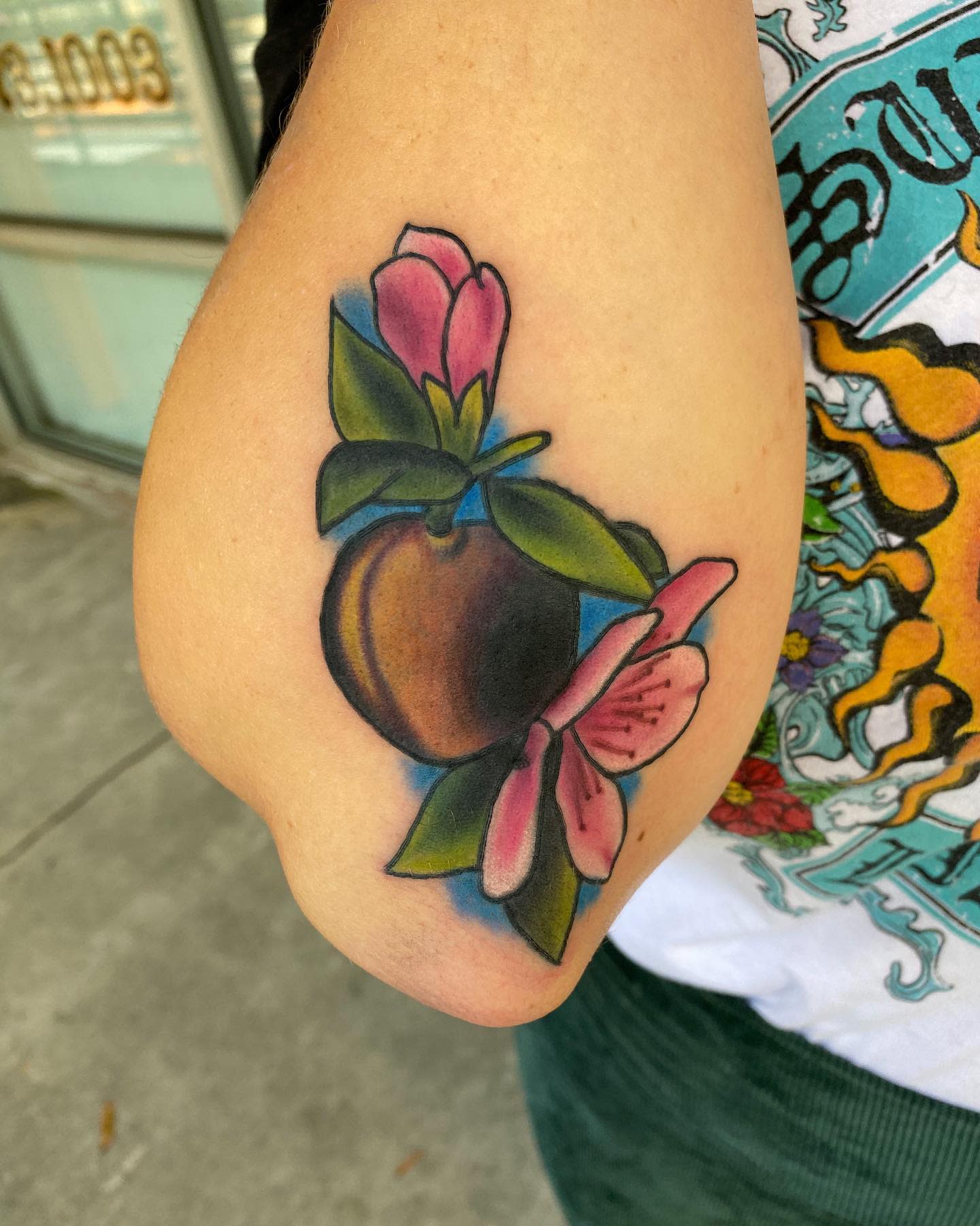Tattoo of a peach with pink flowers and green leaves on an arm near the elbow